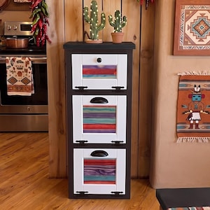 May include: A three-drawer cabinet with white-framed doors and a black top. The drawers display colorful, layered designs. Two decorative cactus figures sit on top. The kitchen setting includes Southwestern-style decor, chili pepper garlands, and a patterned table runner.