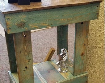 May include: A rustic, handmade wooden side table with a distressed teal finish. The table features a lower shelf and a natural wood top. Decorative candles and a silver figurine are displayed on the table.