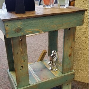 May include: A rustic, handmade wooden side table with a distressed teal finish. The table features a lower shelf and a natural wood top. Decorative candles and a silver figurine are displayed on the table.