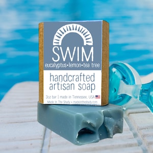 May include: A handcrafted artisan soap bar with the word "SWIM" and the scent "eucalyptus lemon tea tree" printed on the label. The soap is a blue-gray color and is 3oz. The background shows a pool and a pair of goggles.