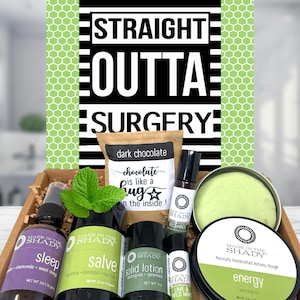 May include: A gift box containing a brown paper pouch labeled dark chocolate, a purple sleep spray, a green salve, a solid lotion, a rollerball, and a tin of light green activity dough. A sign behind the box reads STRAIGHT OUTTA SURGERY.