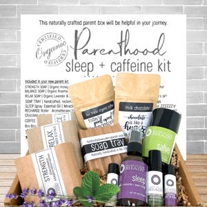 May include: A parenthood sleep and caffeine kit containing various items including bars of soap labeled Strength, Balance, and Relax, a wooden soap tray, a bag of fair trade organic coffee, a bag of milk chocolate, a sleep spray, and essential oil rollers.