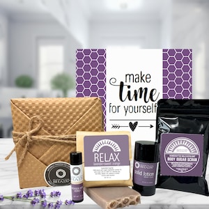 May include: A gift set featuring a variety of self-care products. The set includes a wrapped package, a sign that says "make time for yourself", a bar of soap, a lip balm, a solid lotion, and a bag of body sugar scrub.