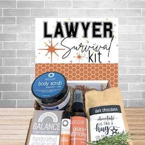 May include: A gift box labeled "Lawyer Survival Kit" contains a body scrub, artisan soap, dark chocolate, and other self-care items. The box includes a body scrub with peppermint and coffee, a bar of soap, and a spray bottle. The dark chocolate is in a brown paper bag.
