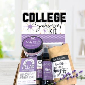 May include: A college survival kit in a cardboard box. The kit includes a body scrub, artisan soap, lip balm, sleep spray, and a bag of milk chocolate. The kit is decorated with lavender and purple accents.