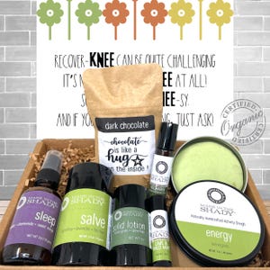 May include: A gift box containing various self-care products. Includes a spray bottle labeled "sleep", a jar of salve, a solid lotion, a dark chocolate bag, and a container of activity dough. The box also contains a small bottle of essential oil and a small container of lime and wild mint.