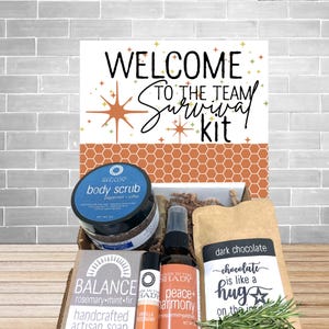 May include: A gift box labeled "Welcome to the Team Survival Kit" includes a body scrub, artisan soap, dark chocolate, and other self-care items. The box features a sign with a starburst design and the words "chocolate is like a hug".