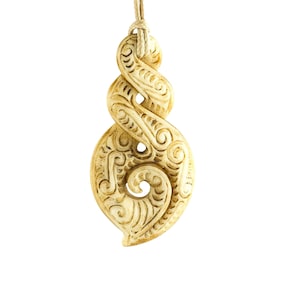 May include: A carved bone pendant with a traditional Maori design. The pendant is a light brown color and has a rope attached to it.