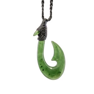 May include: A green jade fish hook pendant on a black cord necklace. The hook is carved in the shape of a traditional Maori fish hook.