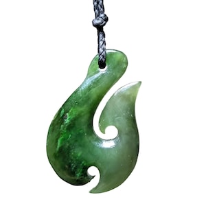 May include: A green jade pendant in the shape of a fish hook, suspended from a black cord. The pendant has a smooth, polished surface with variations in color, ranging from light to dark green. The hook design is a traditional symbol.
