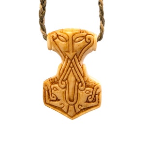 May include: Cream-colored pendant in the shape of Thor's hammer, with intricate carved details. The pendant is suspended from a braided cord. The hammer design features stylized animal heads and geometric patterns, showcasing a Viking aesthetic.