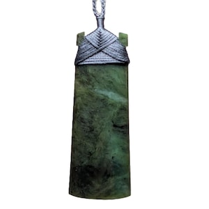 May include: A rectangular pendant crafted from mottled dark green stone, featuring a smooth, polished surface with natural black inclusions. The top is secured with a decorative, intricate silver-coloured cord wrapping that forms a loop for suspension.