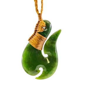 May include: A green jade Maori fish hook pendant with a brown cord. The hook is a traditional symbol of good luck and prosperity.
