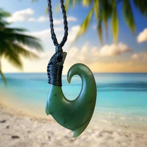 May include: A green jade pendant in the shape of a fish hook, suspended from a black braided cord. The hook is carved with smooth curves and a polished finish. The background shows a beach scene with palm trees and ocean.