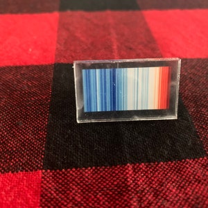 May include: A rectangular, clear acrylic pin featuring a colorful gradient design. The gradient transitions from blue to white to red, resembling a temperature scale. The pin is set against a red and black plaid background.