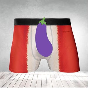 Puede incluir: Bóxers con un diseño rojo y blanco, con un gráfico de berenjena morada. Los bóxers tienen una cintura negra y una zona de bolsa blanca. El fondo es una pared gris claro y un suelo de madera.