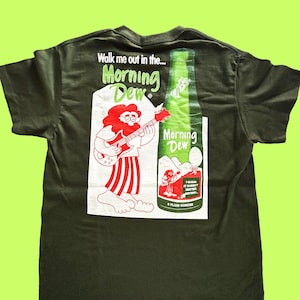 May include: Green t-shirt with a vintage-style graphic of a cartoon character playing a guitar and a bottle of Morning Dew soda. The text on the shirt reads "Walk me out in the... Morning Dew".