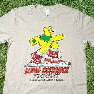 May include: A beige t-shirt with a cartoon illustration of a yellow bear wearing a green leaf crown and red sneakers. The bear is running on a mountain with flames. The text "LONG DISTANCE runner Get up. Get out. Get out of the door." is printed below the bear.