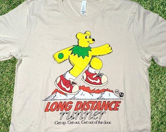 May include: A beige t-shirt featuring a cartoon illustration of a yellow bear wearing a green leaf crown and red trainers. The bear is running on a mountain with flames. The text "LONG DISTANCE runner Get up. Get out. Get out of the door." is printed below the bear.