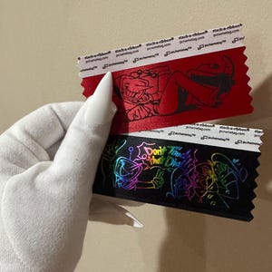 May include: Two decorative ribbons are held up. The top ribbon is red with a black illustration and the text "pcname tag.com". The bottom ribbon is black with a colorful illustration and the text "Don't Touch That Dial".