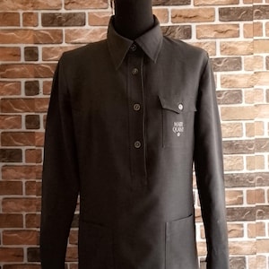 May include: A dark grey long-sleeved dress shirt with a collar, buttoned front, and two pockets. The left chest pocket features the text "MARY QUANT". The shirt is displayed on a mannequin.