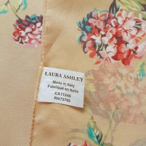 Op de afbeelding: Zijden sjaal met een bloemmotief in rood, roze en teal op een lichtperzik achtergrond. Het label luidt "LAURA ASHLEY Made in Italy".