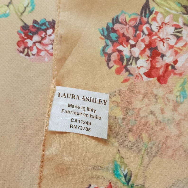 Op de afbeelding: Zijden sjaal met een bloemmotief in rood, roze en teal op een lichtperzik achtergrond. Het label luidt "LAURA ASHLEY Made in Italy".