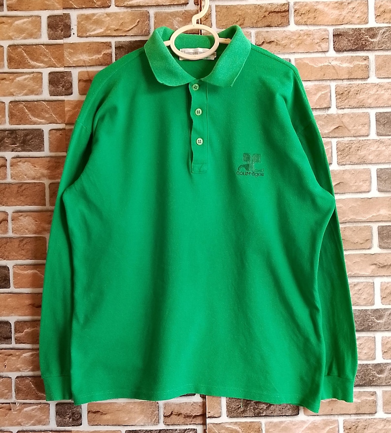 May include: A long-sleeved, bright green polo shirt with a collar and three buttons. The shirt has a small logo embroidered on the chest. The shirt is hanging on a white hanger against a brick wall.