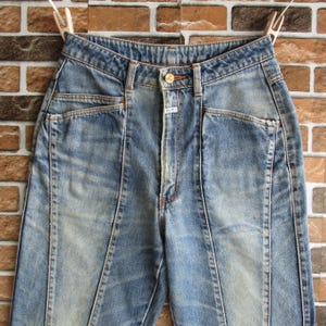 May include: Pale blue denim jeans with a high waist and a button fastening. The jeans feature front pockets and a distinctive seam design down the front of each leg. The jeans are hanging on a clothesline with a brick background.