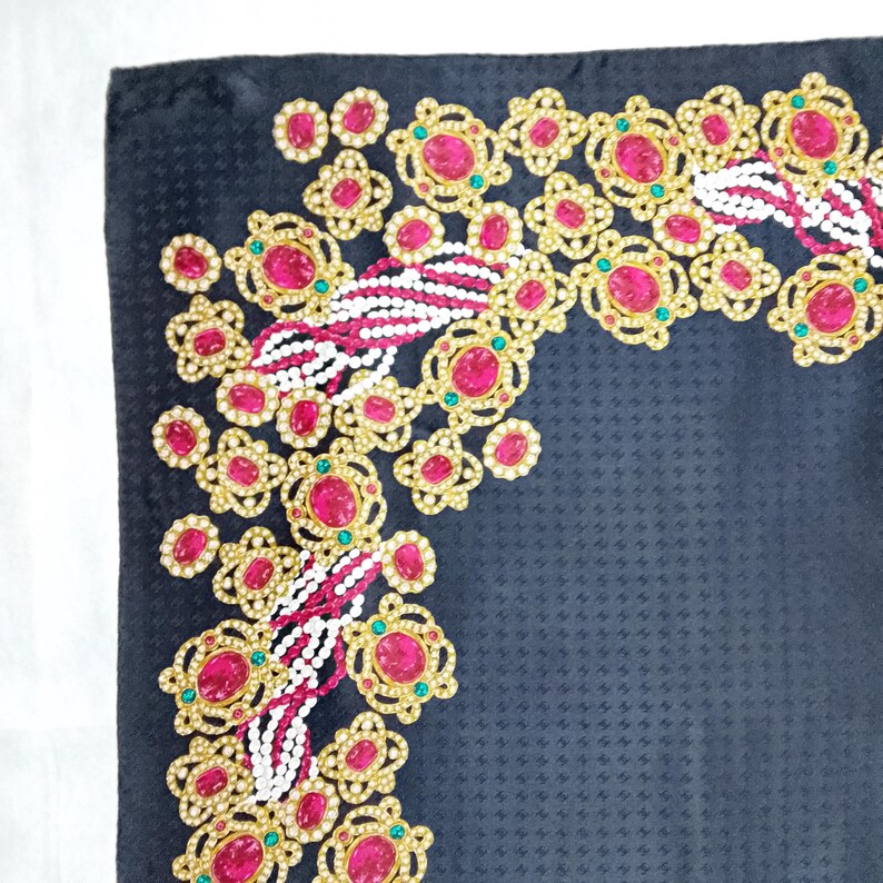 May include: A dark navy blue textile with an ornate, jeweled border. The border features gold-toned filigree, large pink gemstones, small green accents, and strands of white and pink beads. The fabric has a subtle, textured pattern.