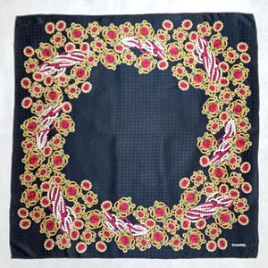 May include: A square, black silk scarf with a repeating floral pattern in gold, red, and white. The scarf has a textured surface and the word "CHANEL" is printed in white in the bottom right corner.