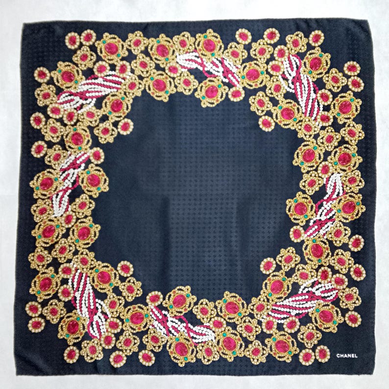May include: A square, black silk scarf with a repeating floral pattern in gold, red, and white. The scarf has a textured surface and the word "CHANEL" is printed in white in the bottom right corner.