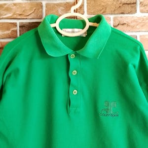 May include: A bright green polo shirt with a classic collar and three-button placket. The shirt features a small embroidered logo on the chest and is made of a textured fabric. The shirt is hanging on a white hanger.