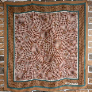 May include: A brown silk scarf with a geometric pattern of squares and rectangles in shades of brown, white, and teal. The scarf has a teal border with a repeating pattern of a stylized "G". The text "PIERRE BALMAIN" is printed in white on the bottom right corner of the scarf.