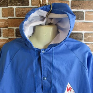 Pode incluir: Uma jaqueta azul com capuz e um logotipo de galo vermelho. A jaqueta tem fecho de correr e capuz com cord&atilde;o.