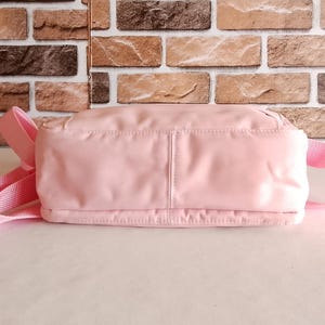 Puede incluir: Un bolso de hombro rosa claro con una correa a juego. El bolso es rectangular con una textura suave y ligeramente brillante. El fondo presenta una pared de ladrillos.