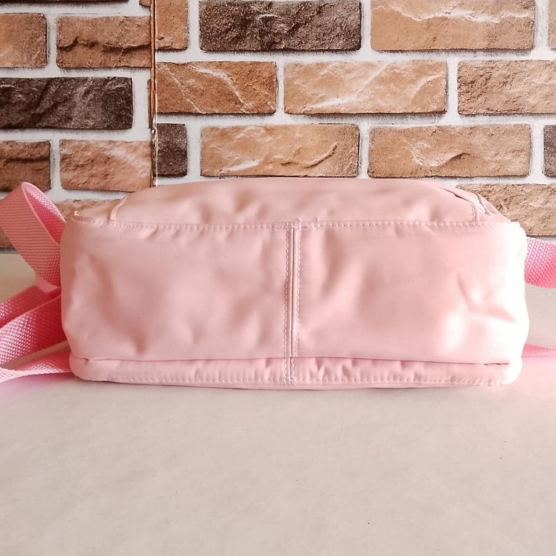 Puede incluir: Un bolso de hombro rosa claro con una correa a juego. El bolso es rectangular con una textura suave y ligeramente brillante. El fondo presenta una pared de ladrillos.