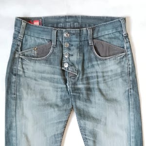 May include: Faded blue denim jeans with a button fly and decorative buttons. The jeans feature grey pocket accents and a red tag on the waistband. The denim has a worn, distressed appearance.