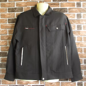 Peut inclure: Une veste de travail noire avec une fermeture éclair, deux poches et des détails de passepoil blancs. La veste a des détails de couture rouge et un petit logo sur la poitrine gauche.