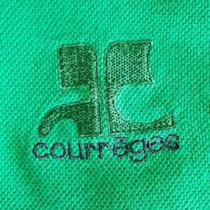 May include: Close-up of a green polo shirt with an embroidered logo. The logo is a stylized design with the word "courr&egrave;ges" embroidered in black below it. The shirt has a textured surface.