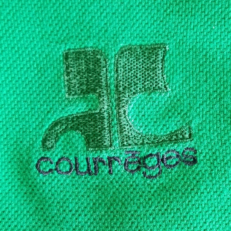 May include: Close-up of a green polo shirt with an embroidered logo. The logo is a stylized design with the word "courr&egrave;ges" embroidered in black below it. The shirt has a textured surface.