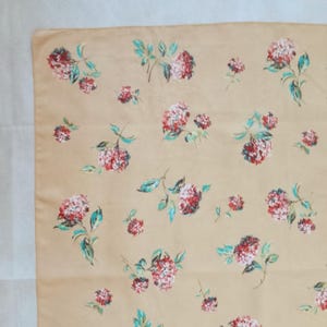 Op de afbeelding: Een beige stof met een herhaald bloemenpatroon. De bloemen zijn rood en roze met groene bladeren. De stof heeft een zacht, licht gekreukt uiterlijk.