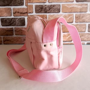 Puede incluir: Un bolso bandolera rosa claro con una correa ajustable a juego. El bolso tiene un compartimento principal con cremallera y un bolsillo frontal m&aacute;s peque&ntilde;o. La correa es de material tejido y est&aacute; sujeta con herrajes met&aacute;licos plateados.