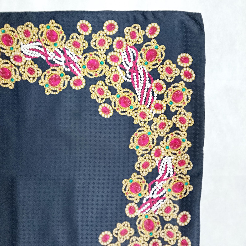 May include: A dark navy blue scarf with a repeating pattern. The scarf's border features a decorative design with gold-colored filigree, pink gemstones, white beads, and small green accents. The design is concentrated in the corners.