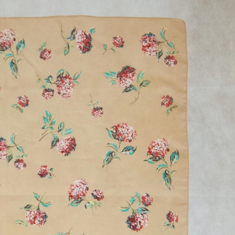 Op de afbeelding: Vierkante beige stof met een herhalend bloemenpatroon. De bloemen zijn rood en roze, met groene en blauwgroene bladeren. Het kan een zakdoek of sjaal zijn.