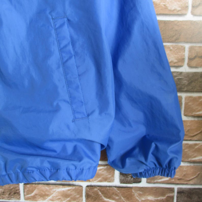 Pode incluir: Uma jaqueta de nylon azul com fecho de correr e um bolso no lado esquerdo. A jaqueta tem um cord&atilde;o na parte inferior e punhos el&aacute;sticos.