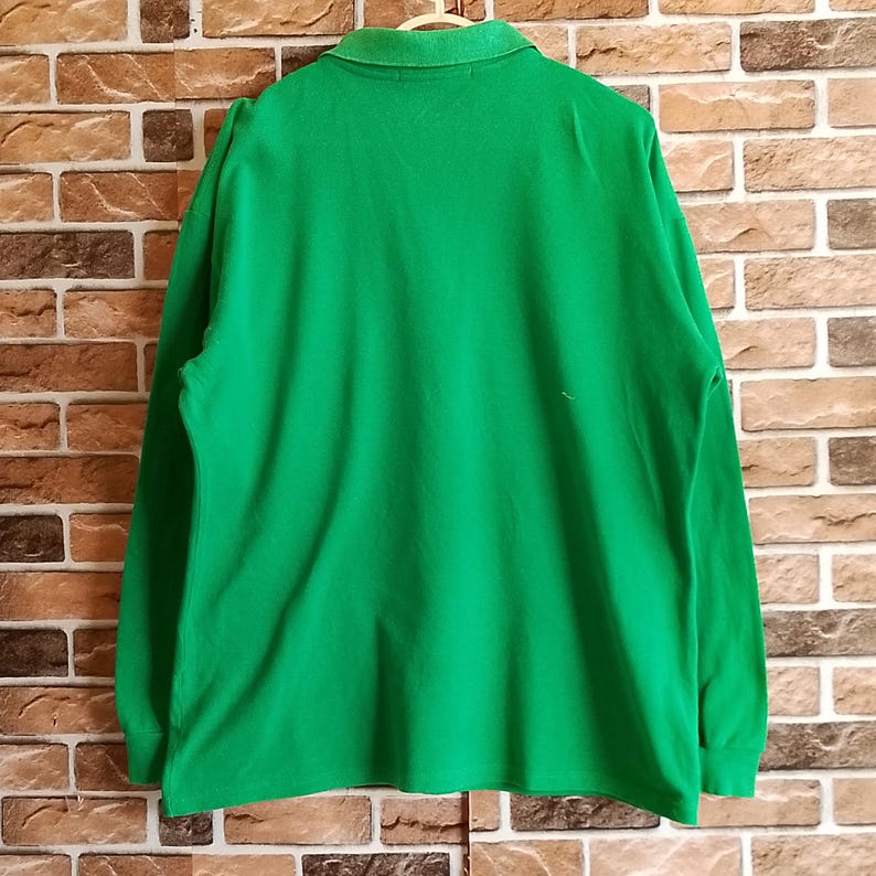 May include: A bright green long-sleeved polo shirt. The shirt has a collared neckline and is made of a soft, likely cotton material. The shirt is hanging on a wooden hanger against a brick wall background.