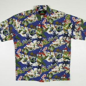 Può includere: Una camicia blu a maniche corte con una stampa tropicale che presenta ananas, fiori, palme e uccelli. La camicia ha un colletto abbottonato e un'etichetta verde con il testo "Island Wear" sul lato sinistro del colletto.