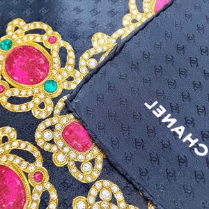 May include: A navy blue silk scarf with a repeating pattern of interlocking Cs. The scarf features ornate gold and rhinestone designs with large pink and green gemstones. The word "CHANEL" is printed in white.