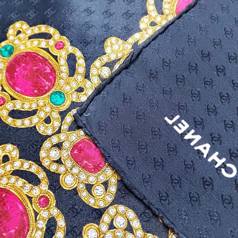 May include: A navy blue silk scarf with a repeating pattern of interlocking Cs. The scarf features ornate gold and rhinestone designs with large pink and green gemstones. The word "CHANEL" is printed in white.